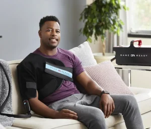 Mężczyzna używa urządzenia Game Ready na ramieniu, siedząc na kanapie w domowym salonie, obok stolika z urządzeniem Game Ready.