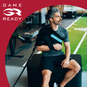 Mężczyzna siedzący na ławce, korzystający z urządzenia Game Ready, które jest zamocowane na jego ramieniu. W tle widoczna jest siłownia, a logo Game Ready znajduje się w lewym górnym rogu.