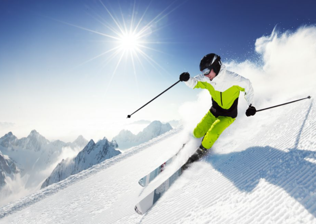 Narciarz w jasnym stroju zjeżdżający ze stoku na nartach w słoneczny zimowy dzień, otoczony majestatycznymi górami.