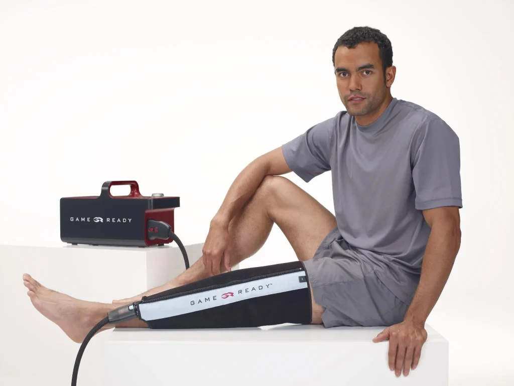 Mężczyzna siedzący na białym podwyższeniu z mankietem Game Ready na kontuzjowanym kolanie.
